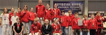 Drużyna sekcji karate Raion Fight Club w klubowych bluzach, część zawodników z medalami na szyi, wspólne zdjęcie po zawodach na hali sportowej